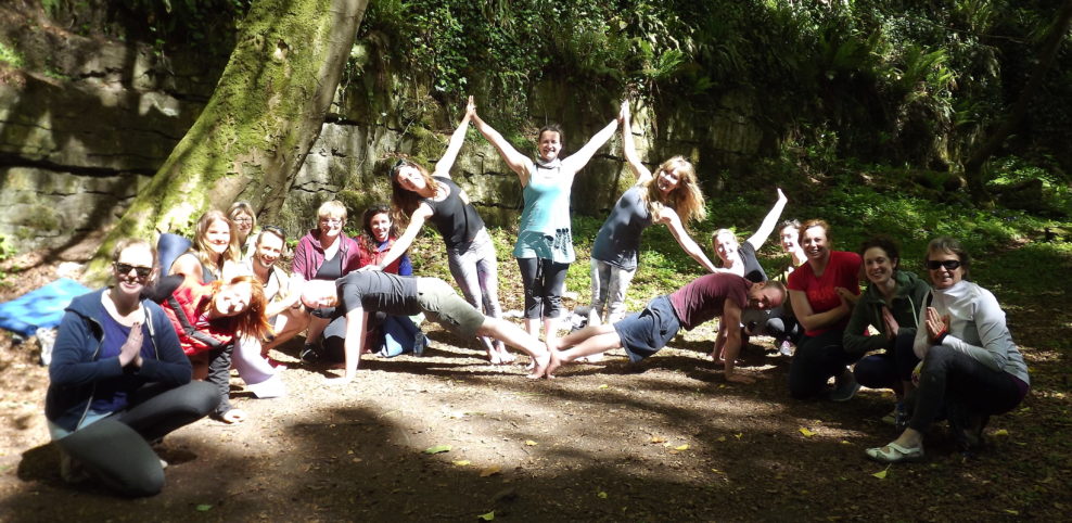 A group of people outdoors in a wooded area, forming a human pyramid and other playful poses, smiling and waving at the camera. Sunlight filters through the trees, creating a cheerful, lively atmosphere.