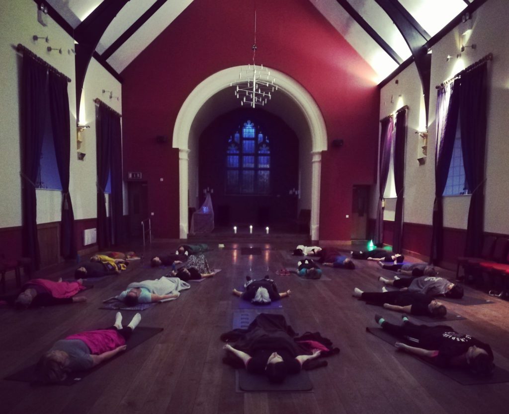A group of people lie on yoga mats in a large, dimly lit hall with high vaulted ceilings, arched windows, and candles at the front—creating a calm, meditative atmosphere for a CorePower Yoga Sligo adventure.