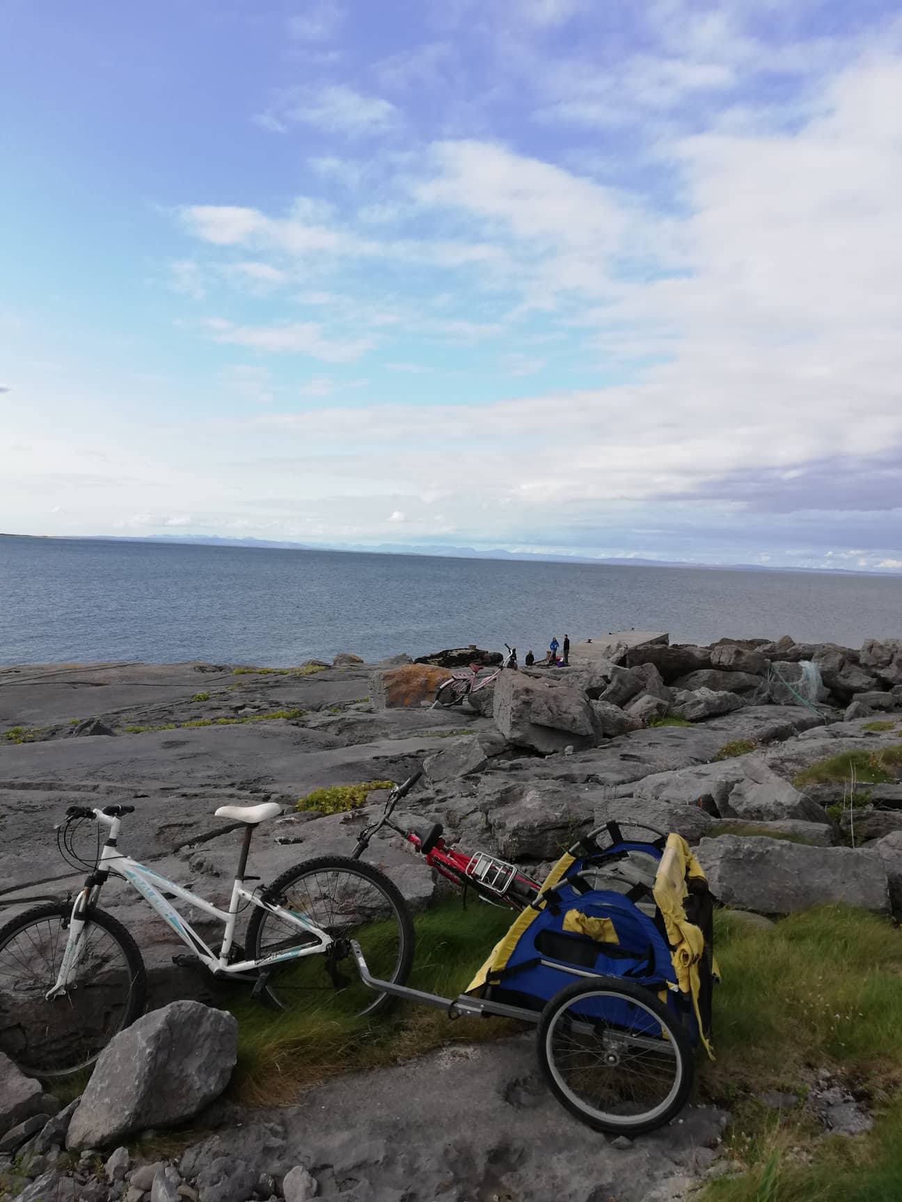 A white bicycle with a blue and yellow trailer stands on rocky ground near the shore, close to Sweathouse, with a calm sea and distant people on the rocks under a partly cloudy sky.