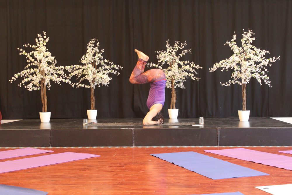 A person in purple clothing performs a headstand on a stage decorated with four potted artificial trees in bloom, with yoga mats laid out on the wooden floor in front, showcasing the spirit of yoga wellness Sligo.