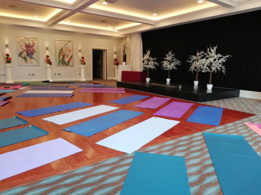A spacious, well-lit room at Sweathouse with colorful yoga mats neatly arranged on the wooden floor, facing a small stage decorated with white artificial trees and floral artwork on the walls.
