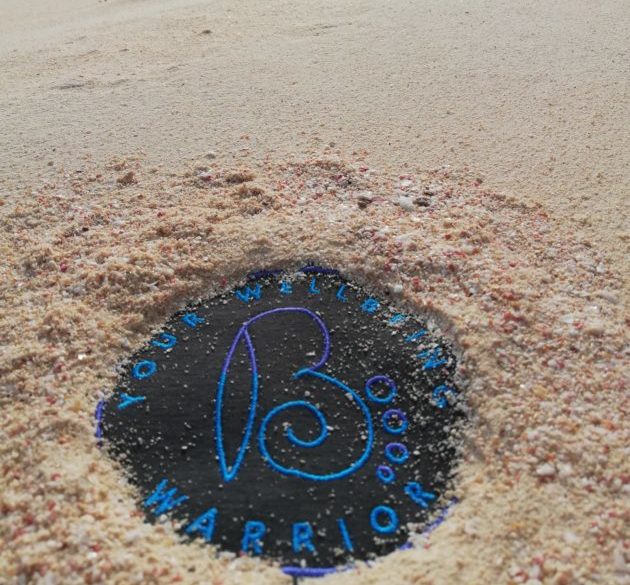 A round object with the words YOUR WELCOMING WARRIOR, partially buried in sand on a Sligo beach. The blue writing surrounds a large stylized B in the center, hinting at a wellbeing adventure along the shore.