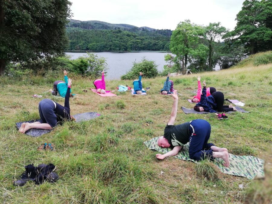 A group of people practice yoga outdoors on mats in a grassy area near a lake, surrounded by trees and hills, with cloudy skies overhead.