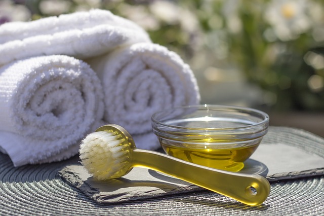 A small glass bowl of oil, a yellow facial brush, and two rolled white towels are arranged on a mat, creating a relaxing spa-like atmosphere outdoors—perfect for sparking the relaxation response before an ayurvedic massage or yoga session.