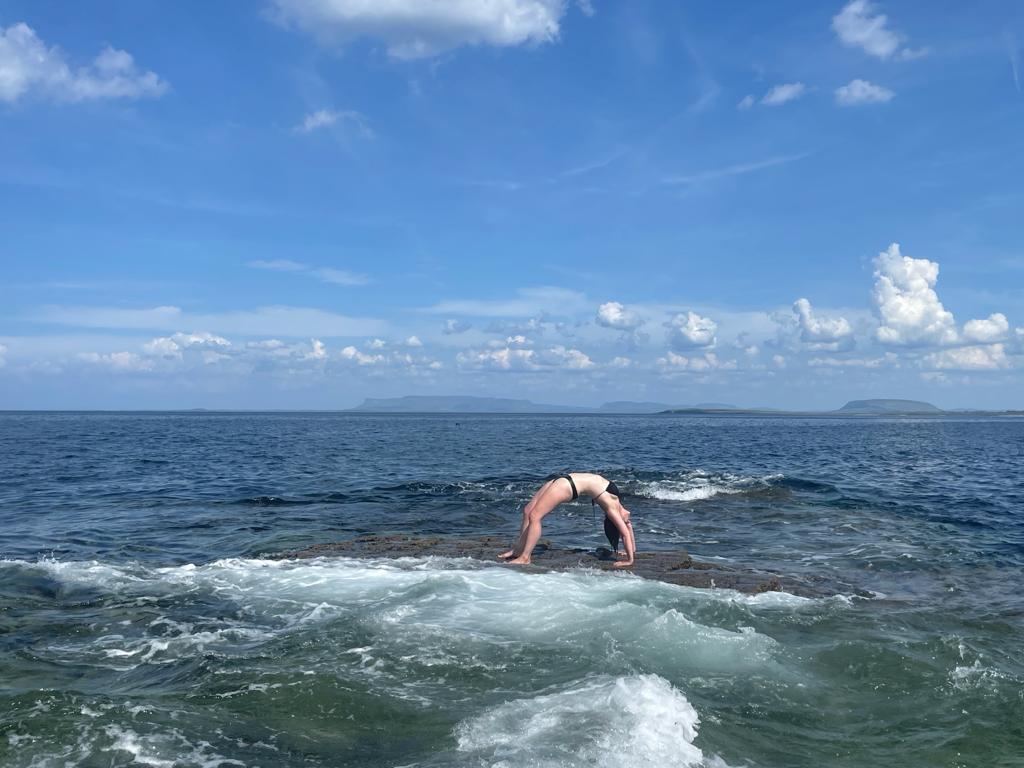 A person in a swimsuit with bold patterns does a backbend on a flat rock in the sea, waves splashing around, under a blue sky with scattered clouds and distant land visible on the horizon.