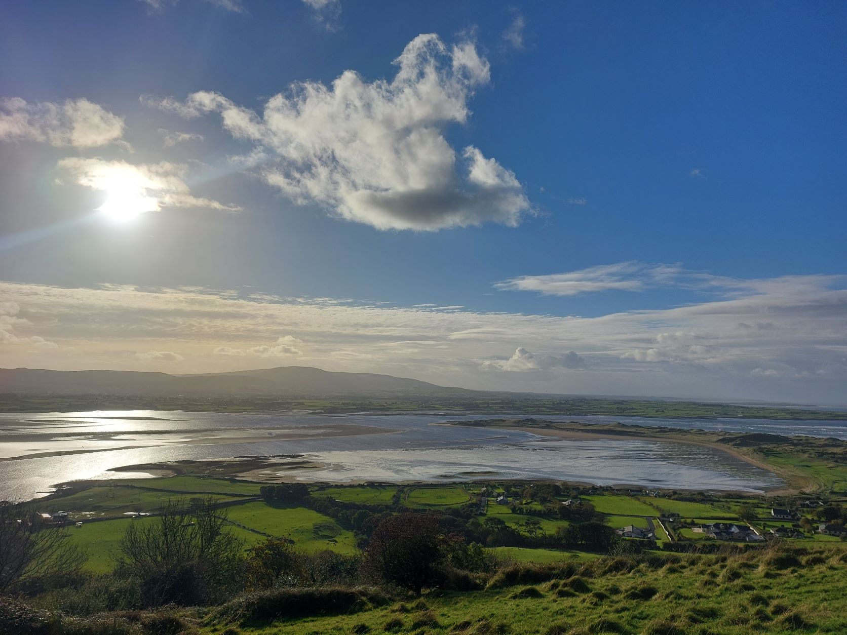 A scenic view of a coastal landscape with green fields, small houses, and a river or inlet stretches beneath a partly cloudy sky. Sunlight bathes the hills in the distance, inviting peaceful afterthoughts amid nature’s quiet beauty.