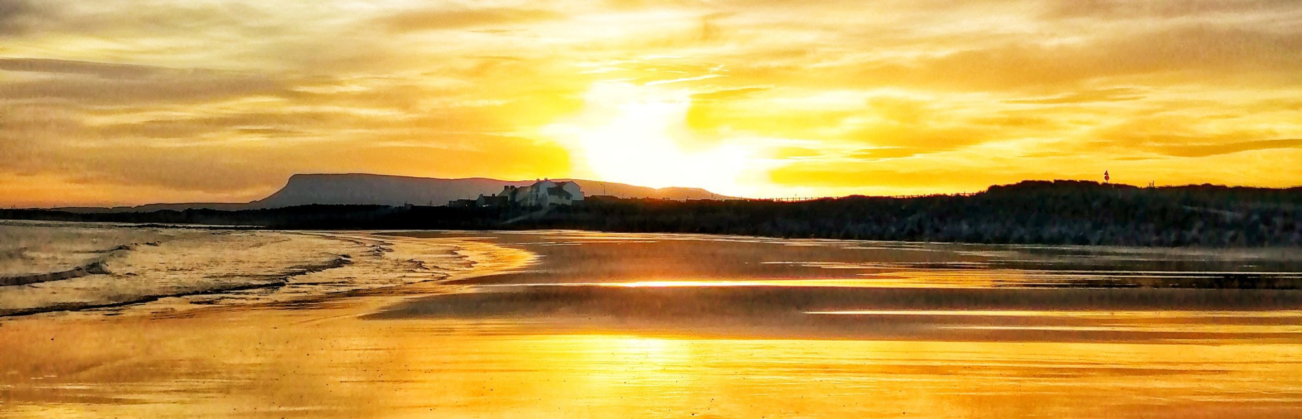 Golden sunset over a sandy beach with the sun low on the horizon, reflecting on wet sand. Silhouettes of distant hills and a building are visible against a glowing, colorful sky.