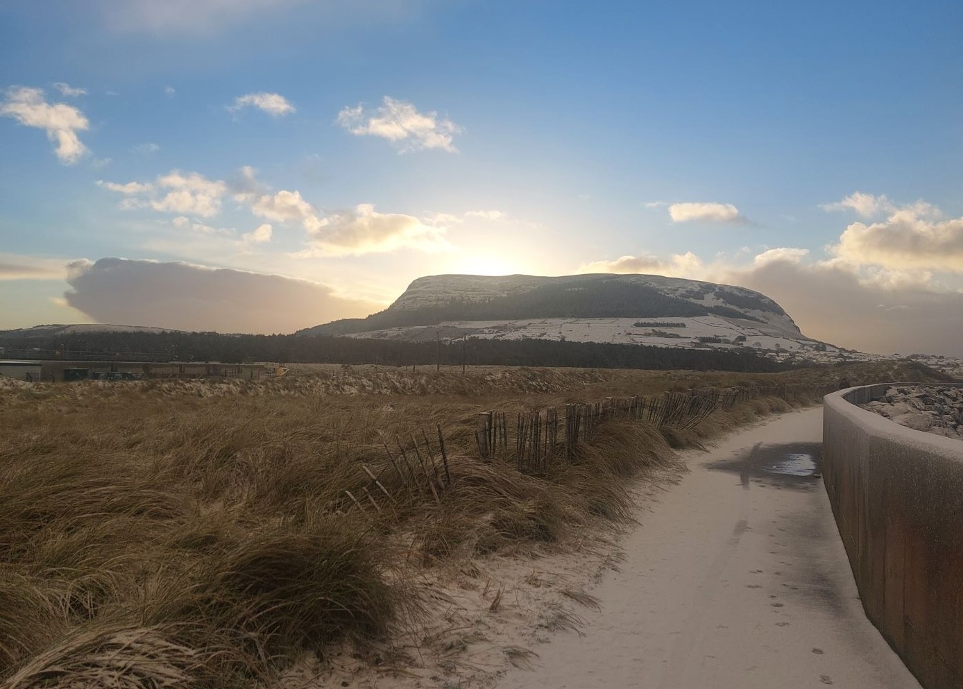 A sandy path lined with tall grass invites reflection—its Time to Plan—as it leads toward a snow-covered hill beneath a partly cloudy blue sky, the sun rising or setting behind the hill.