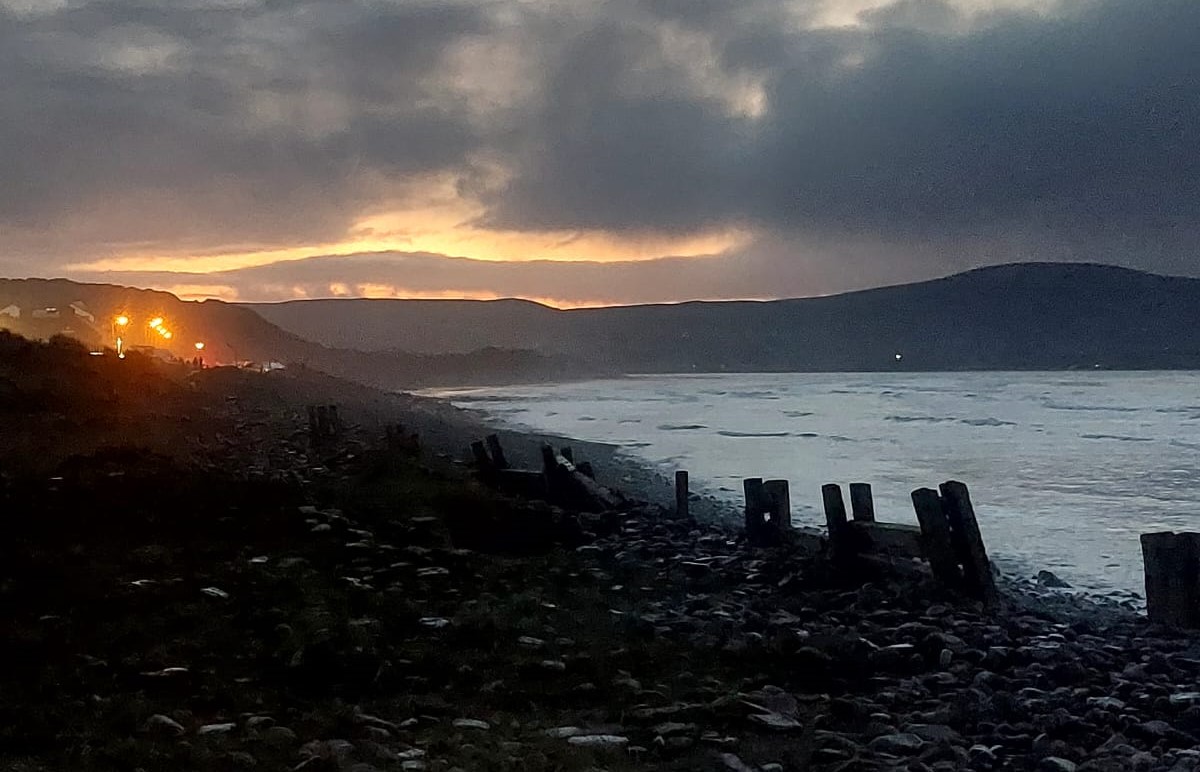 Rocky shoreline at dusk with wooden posts along the coast, distant hills silhouetted against a cloudy sky, and warm lights from buildings glowing on the left—a tranquil setting that invites reflection and understanding sleeps natural rhythms.