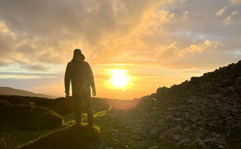 A person stands on rocky terrain at sunset, silhouetted against a bright sky during a solstice hike. Rolling hills and a pile of stones are visible in the background as the sun sinks low on the horizon.