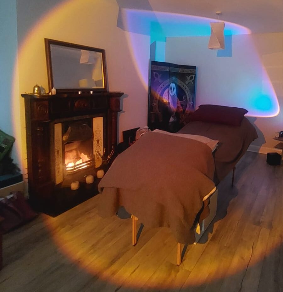 A cozy room with a massage table covered in brown blankets, a lit fireplace with a mirror above, and soft, colorful lights casting a warm glow—perfect for a Reiki Teacher to create a relaxing and healing atmosphere.