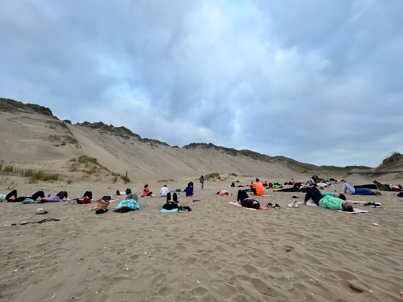 A group of people practice Free Beach Yoga on mats spread across a sandy beach, with large dunes and a cloudy sky in the background.