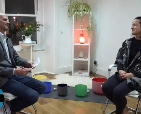 Two people sit facing each other in a cozy, well-lit room with wooden floors. Between them are colorful singing bowls on a mat, a small shelf with a glowing lamp, and plants, creating a calm and relaxed atmosphere.