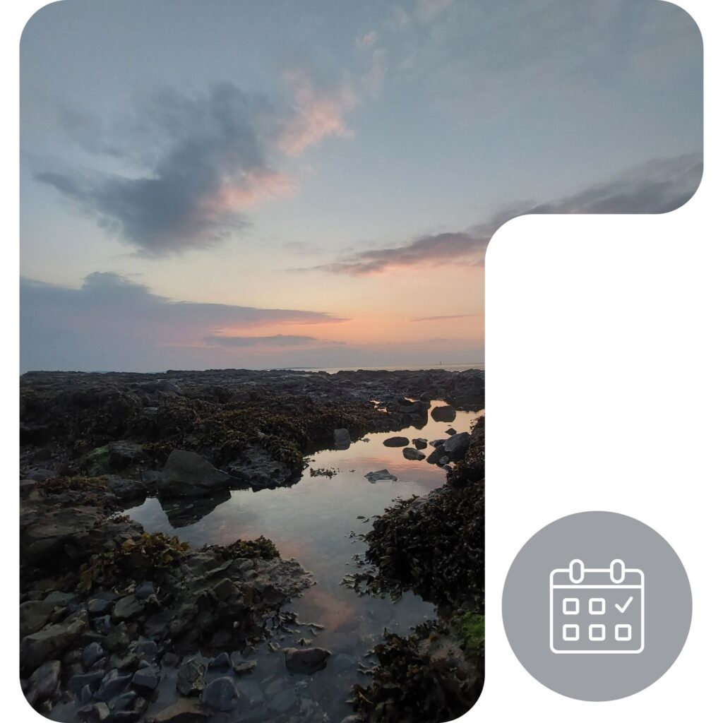 A rocky shoreline with tide pools reflecting a colorful sunset sky, featuring scattered clouds. In the lower right corner, there is a grey calendar icon with a checkmark.