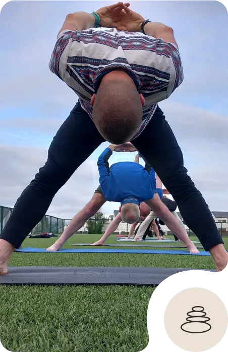 People practicing yoga outdoors on grass, bending forward with legs wide apart and arms clasped behind their backs. The view is from behind, focusing on one person in the foreground. A balance stone icon is in the bottom corner.
