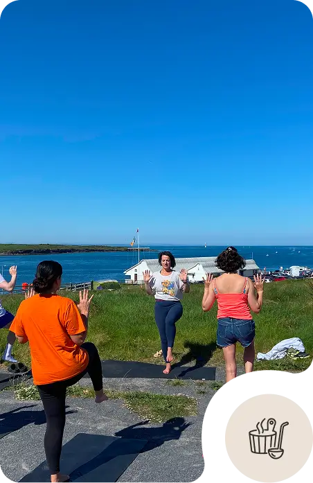 Four people practice yoga outdoors near the water on a sunny day, standing on mats and balancing on one leg with arms raised. A clear blue sky, grassy area, and waterfront buildings are visible in the background.