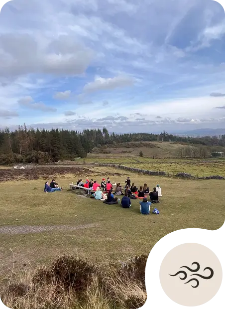 A group of people sit in a circle on a grassy field under a partly cloudy sky, surrounded by trees and rolling hills. An icon with wind symbols appears in the bottom right corner.