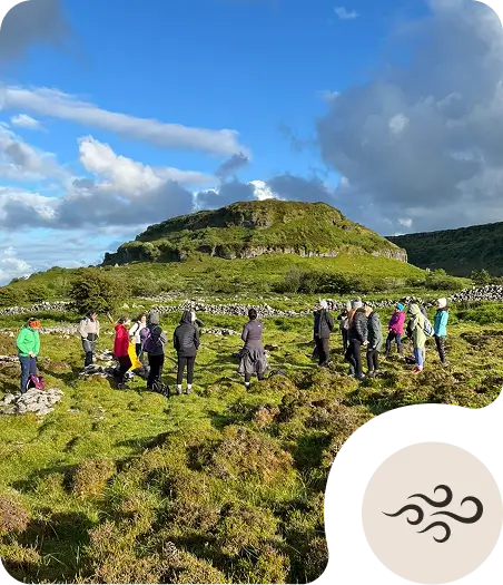 A group of people stand in a grassy, rocky field with a green hill in the background under a blue sky with clouds. The group appears to be gathered for an outdoor activity or tour.