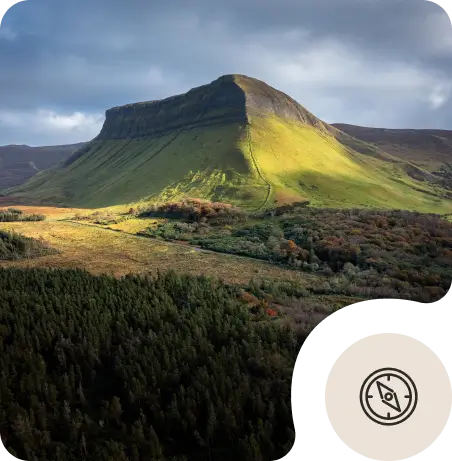 A sunlit, green mountain rises above a patchwork of forests and fields under a cloudy sky. The landscape is vibrant with shadow and light. A compass icon is displayed in the lower right corner.