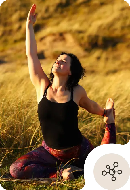 A woman in outdoor exercise clothes practices a yoga pose on grass, raising one arm upward while holding her foot behind her with the other hand. The sunlight highlights her face and the natural background.