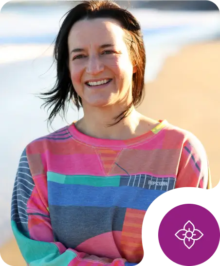 A woman with shoulder-length brown hair smiles on a sunny beach, wearing a colorful patchwork sweatshirt. A purple flower icon appears in the lower right corner.