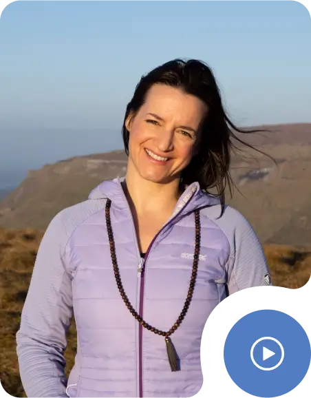 A woman wearing a light purple jacket and beaded necklace smiles outdoors with mountains and clear sky in the background.