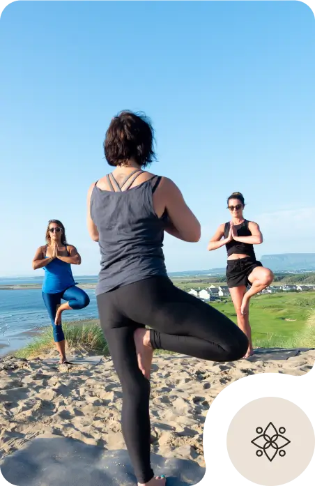 Three women practice yoga outdoors on a sandy hill, standing in a tree pose with hands together. The background shows grassy fields, water, and a blue sky.