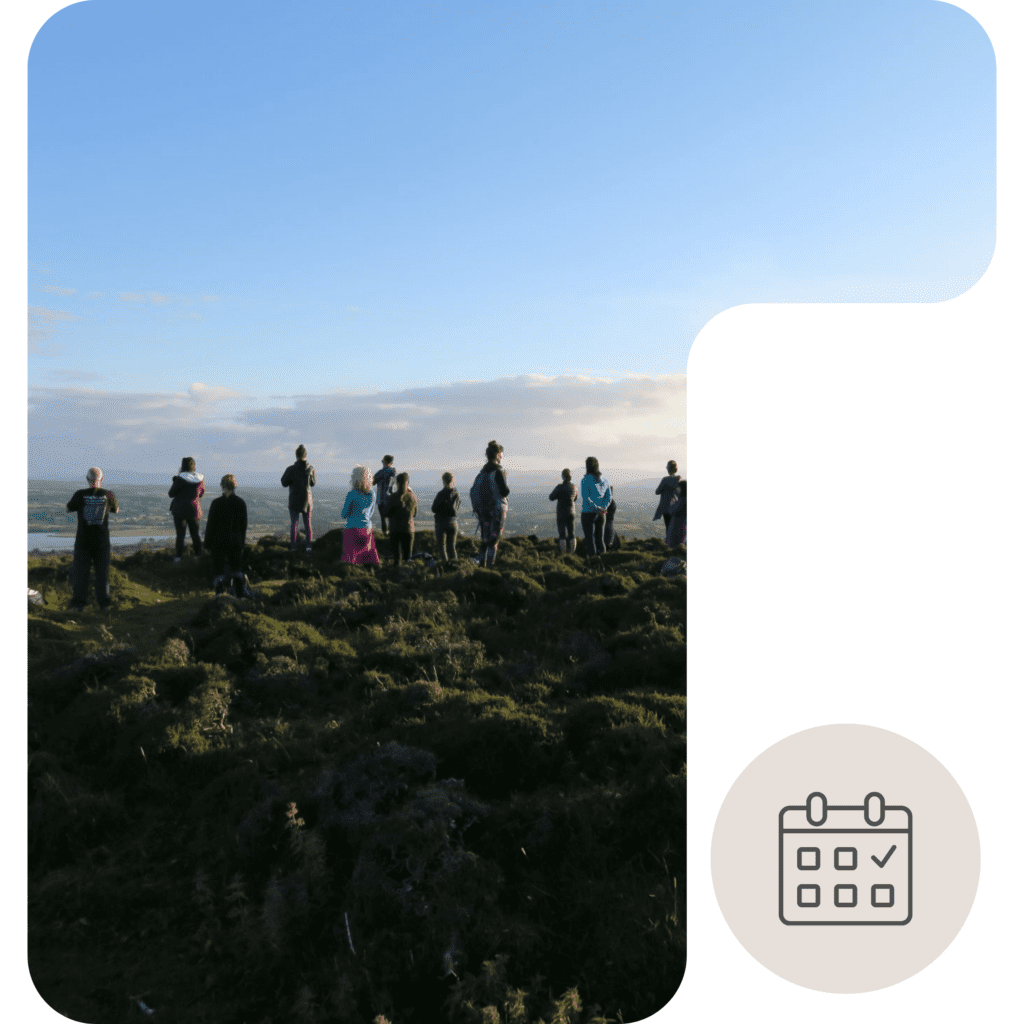 A group of people stands on a grassy hilltop under a clear blue sky, looking out over the landscape. In the bottom right corner, there is an icon of a calendar with a checkmark.
