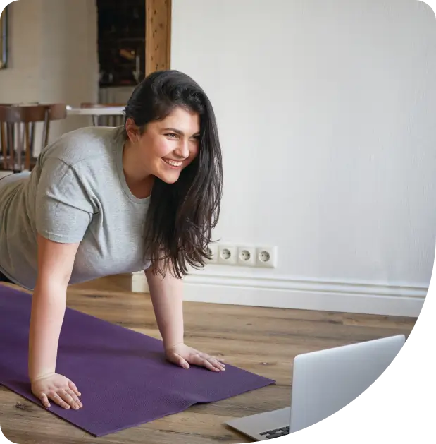 A woman in a gray t-shirt does a plank on a yoga mat indoors, smiling and looking at a laptop in front of her.