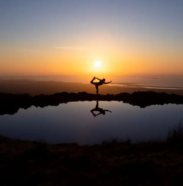 A person practices a yoga pose by a still body of water at sunset, their silhouette and reflection visible against a colorful sky.