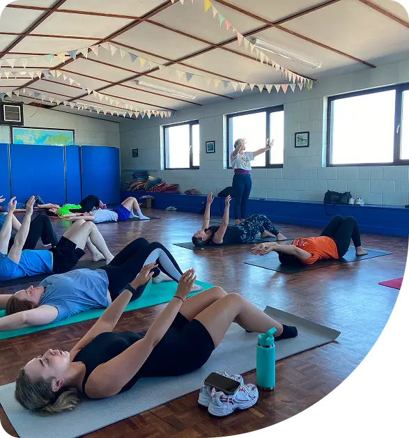 A group of people lie on yoga mats in a bright room with wooden floors, following an instructor who is standing and demonstrating an exercise. Water bottles and small towels are nearby.