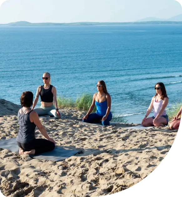 Four women practice yoga while seated on mats on a sandy beach with grass, facing the ocean. The weather is sunny and calm, with blue sky and gentle waves in the background.