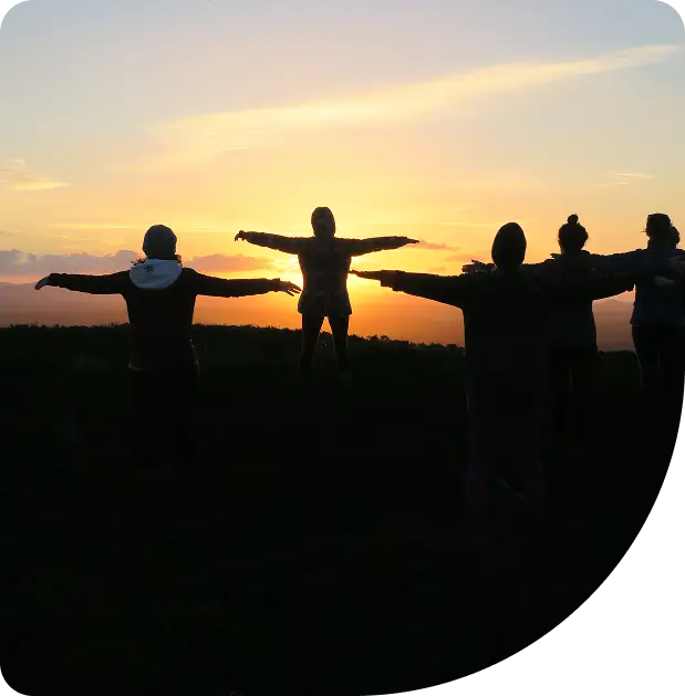 Five people stand with arms outstretched, silhouetted against a sunset sky, creating a peaceful and joyful atmosphere in an open outdoor setting.