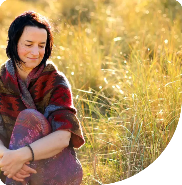 A woman with dark hair, wearing a colorful shawl, sits in a sunlit field of tall grass, smiling gently with her eyes closed and arms resting on her knees.