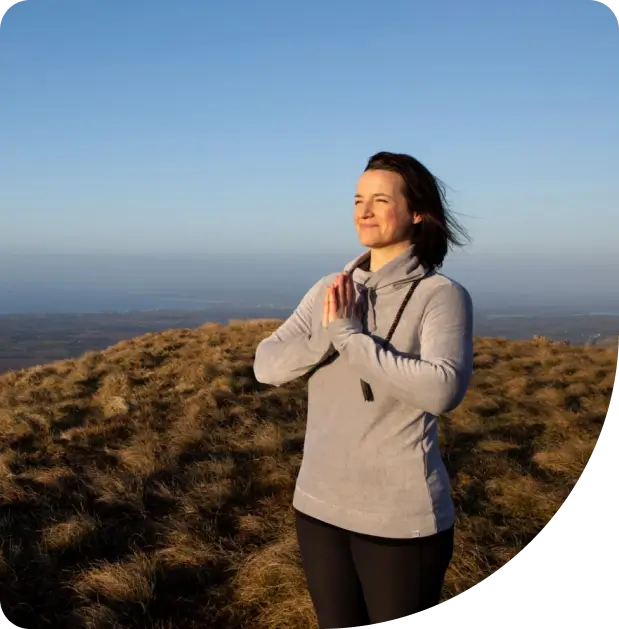 A woman stands outdoors on a grassy hill, eyes closed and hands in a prayer position, enjoying the sunlight and peaceful surroundings under a clear blue sky.