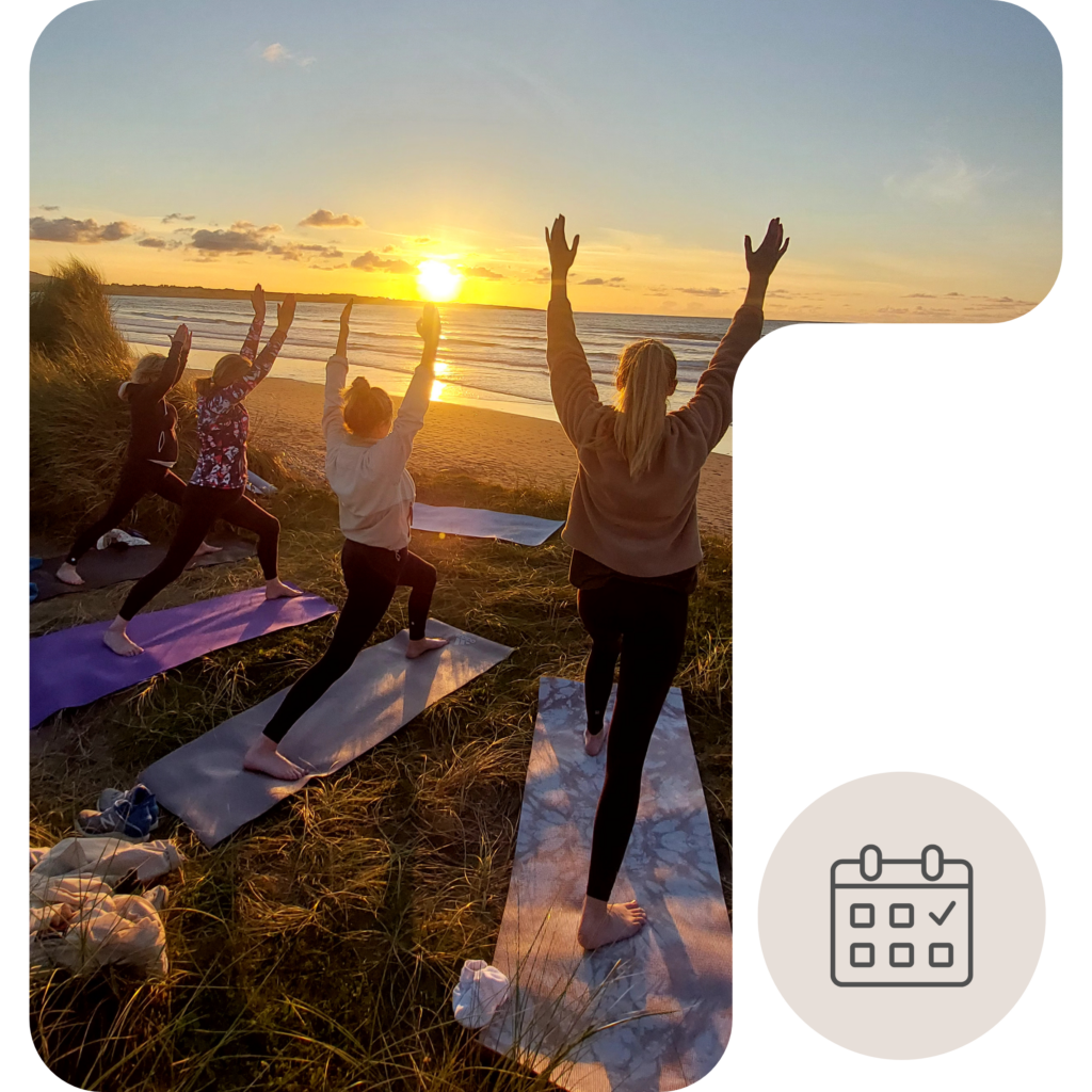 A small group practices yoga on mats among the sand dunes overlooking Strandhill Beach at sunset. With arms raised toward the sky in a standing pose, participants face the golden sun as it reflects across the ocean, creating a peaceful coastal setting bathed in warm evening light.