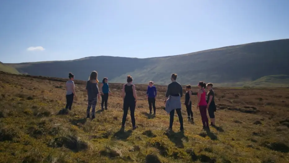 A group of people stand in a circle on grassy moorland under a clear blue sky, with rolling hills in the background.