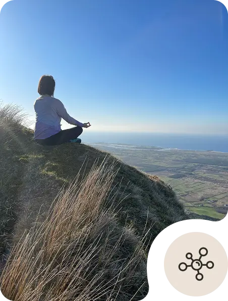 A person sits cross-legged in a meditative pose on a grassy hillside overlooking a vast landscape with fields and the ocean under a clear blue sky. An abstract network icon appears in the bottom right corner.