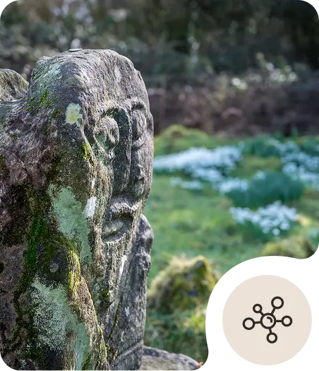 A weathered stone carving with a face stands in a green, mossy outdoor setting, with white flowers in the blurred background. A circular icon with connected dots is overlaid in the bottom right corner.