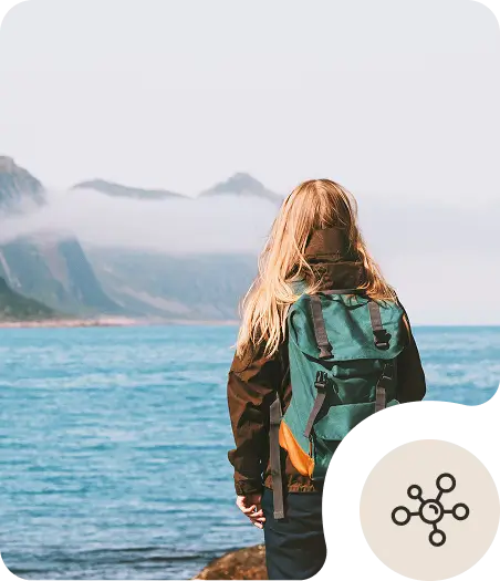 A person with long blonde hair and a green backpack stands by the edge of a calm blue lake, looking toward misty mountains in the distance. An icon with connected dots appears in the lower right corner.
