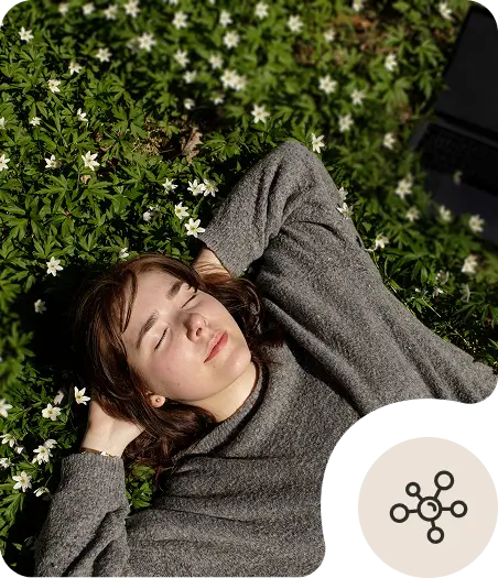 A young woman with brown hair lies in green grass with white flowers, eyes closed and hands behind her head, wearing a gray sweater. A circular icon with a network symbol appears in the bottom right corner.