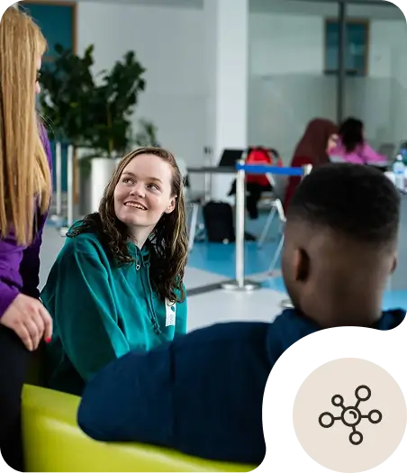 Three people are sitting indoors, engaged in conversation. A woman in a teal hoodie is smiling up at another person, while a third person sits with their back to the camera. There are others in the background.