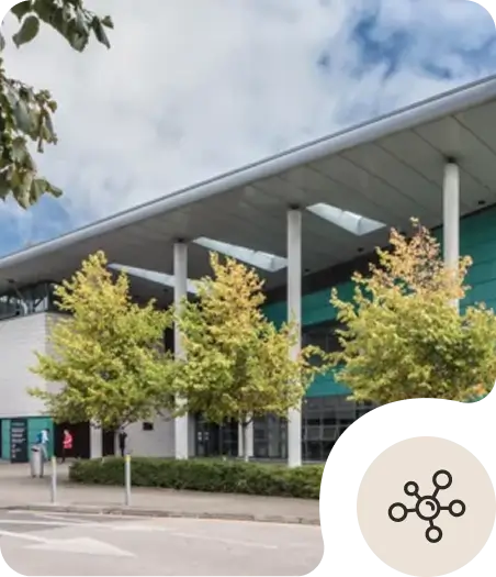 Modern building with a teal and white facade, large windows, and tall white pillars. Trees with green foliage line the front, and a cloudy sky is visible above. A network icon is overlaid in the bottom right corner.
