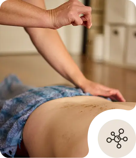 A person sprinkles a substance, possibly herbs or powder, onto someone’s bare back during a massage or body treatment in a calm indoor setting.
