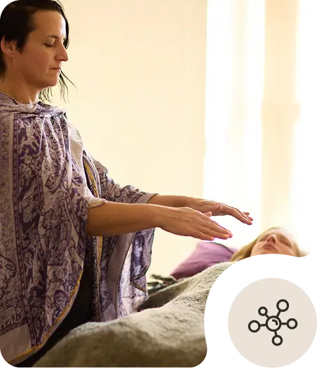 A person stands with hands hovering over another person lying on a table, suggesting a healing or energy therapy session. The person lying down is covered with a blanket.