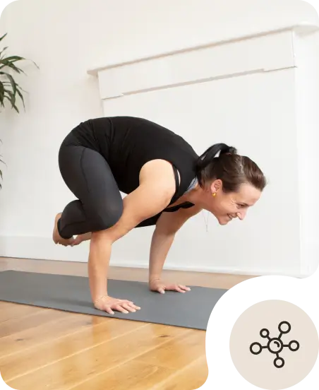 A woman in black athletic wear practices a yoga arm balance pose on a gray mat indoors, smiling and focused, with a plant and white wall in the background.