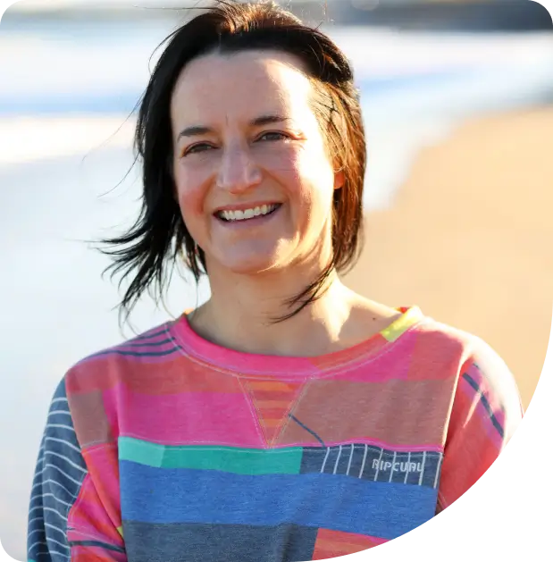 A person with shoulder-length brown hair smiles while standing on a sunny beach, wearing a colorful striped sweatshirt with the Rip Curl logo. The ocean and sand are visible in the blurred background.