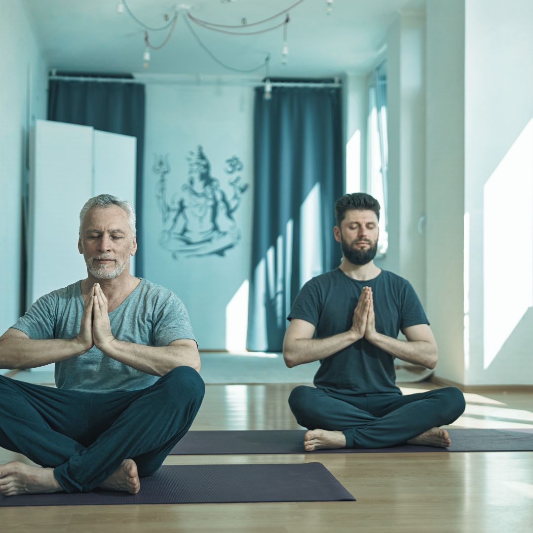Two men practice yoga for men, sitting cross-legged on mats with eyes closed and hands in prayer position, meditating in a bright, serene room adorned with a spiritual wall mural.