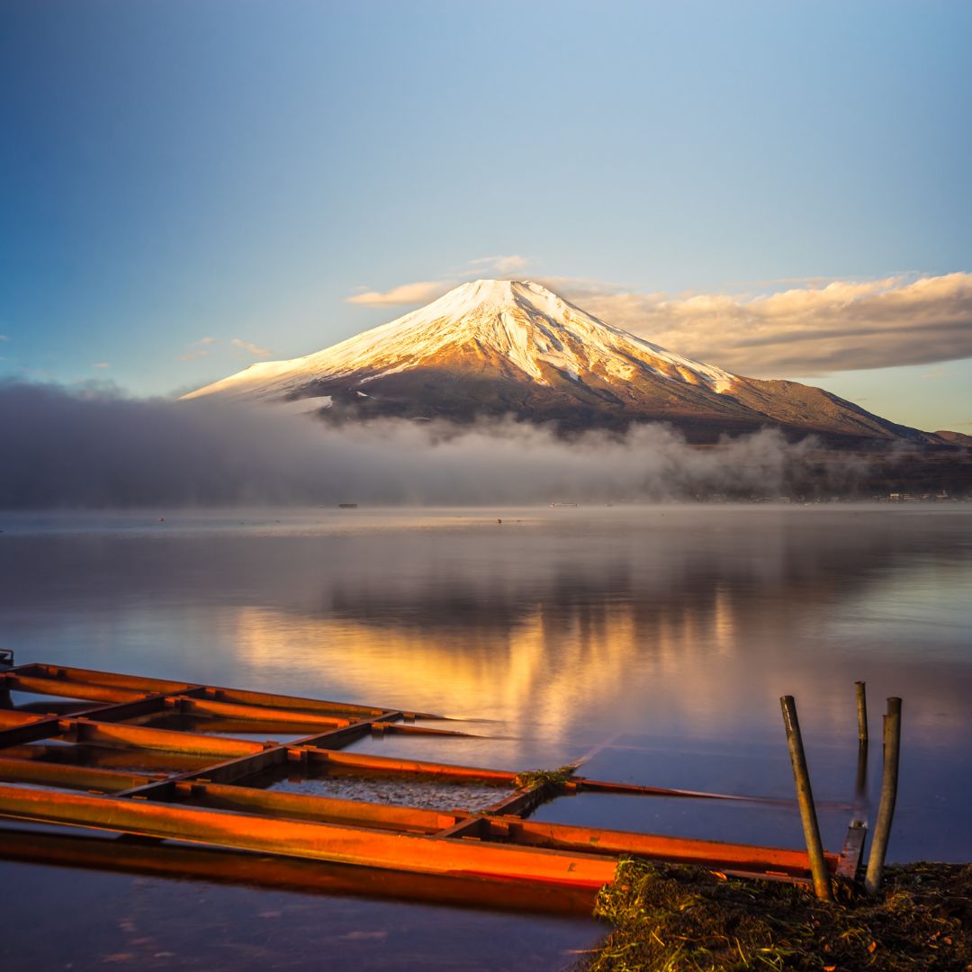 Snow-capped Mount Fuji reflected in a calm lake at sunrise, with orange wooden boats in the foreground and mist rising from the water, evokes the serene spirit of Reiki and echoes traditions of Indigenous Japan.