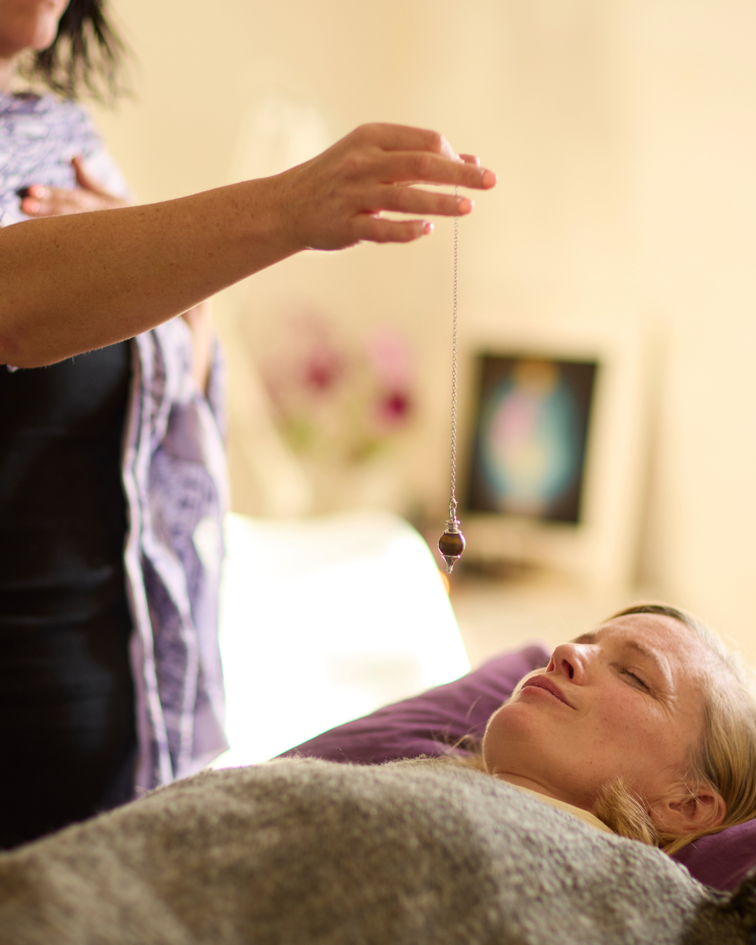 A person holds a pendulum over another lying down with eyes closed, in a relaxed setting, suggesting an energy healing or alternative therapy session inspired by the guidelines of reiki. The background is softly lit and blurred.